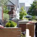 Expat buying agent in the Netherlands handing keys to a client at a wooden gate near a modern residential neighborhood